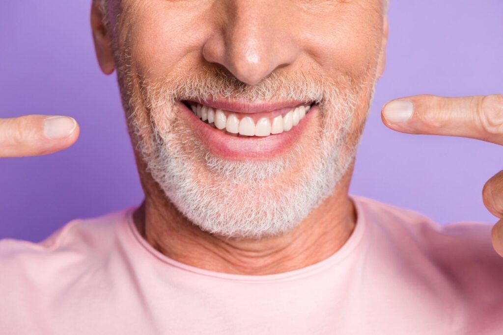 Smiling older man in a pink t-shirt showcasing bright, white teeth against a violet background, emphasizing cosmetic dentistry and smile makeovers.